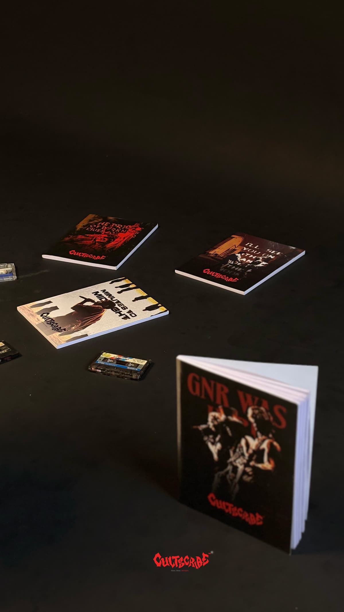CultScribe flat lay — notebooks and cassette tapes on a dark surface with GNR Was Here standing upright.