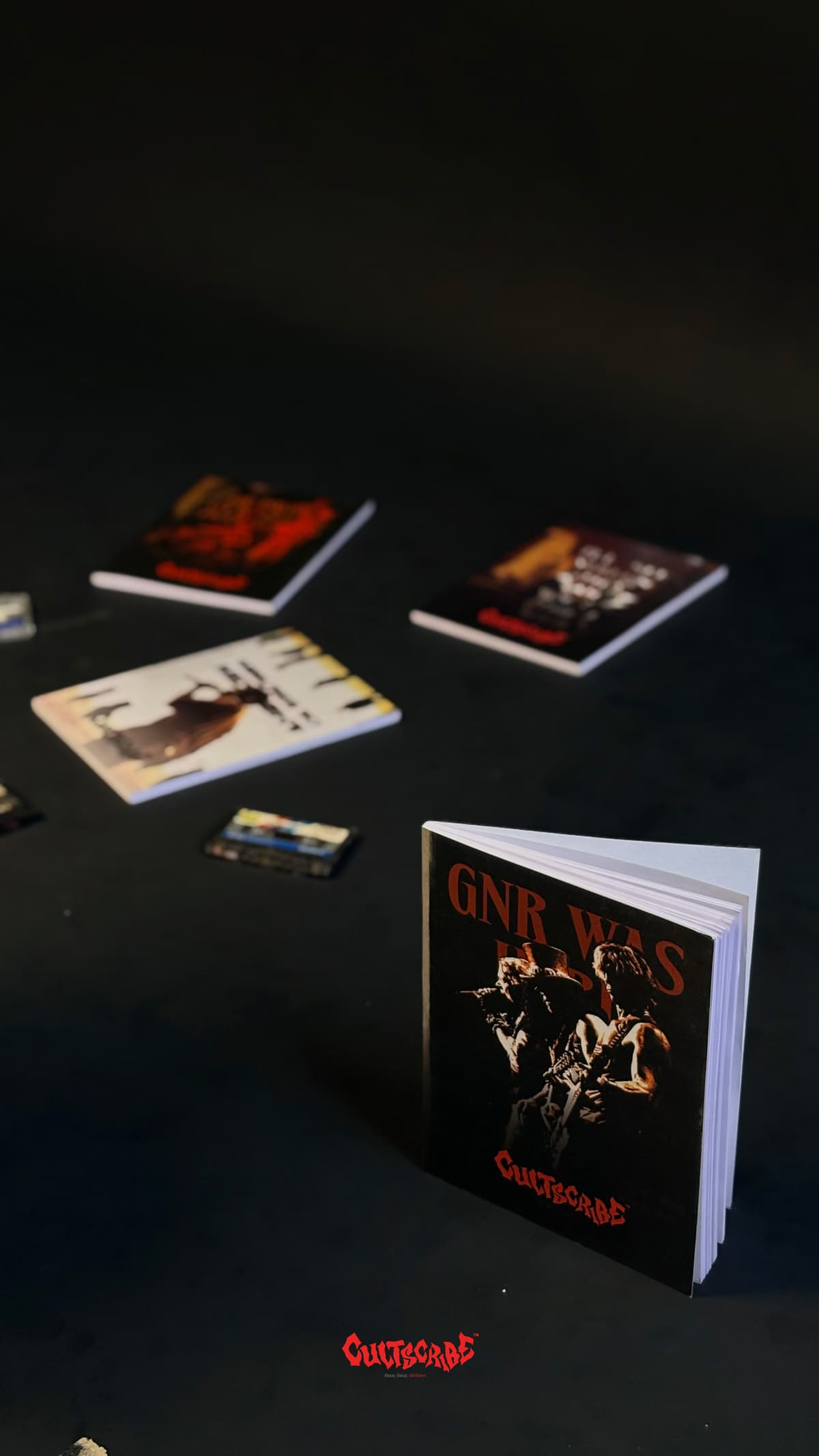CultScribe product lineup — GNR Was Here notebook standing among zines and cassette tapes on a dark surface.